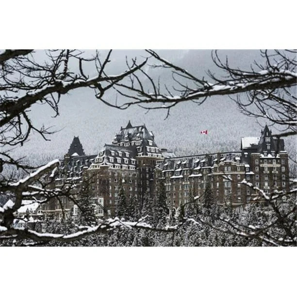 Snow Covered Banff Springs Hotel Framed by Tree Limbs with Snow Covered Trees On Mountain Hillside in The Background - Ba Poster Print by Michael Interisano, 19 x 12